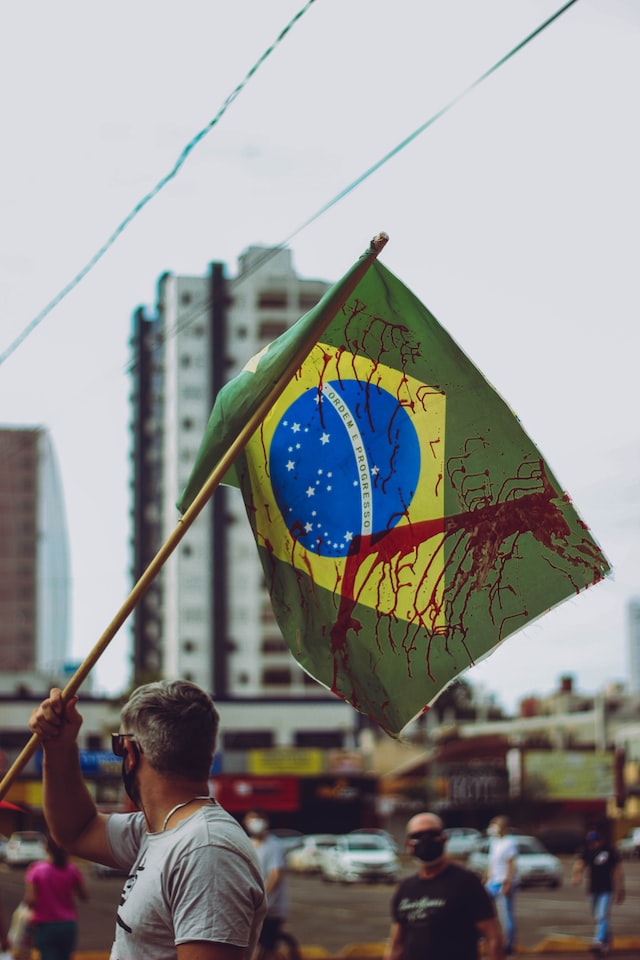 Homem de máscara e camisa cinza de costas, em meio à uma manifestação, segurando uma bandeira do brasil ensanguentada 