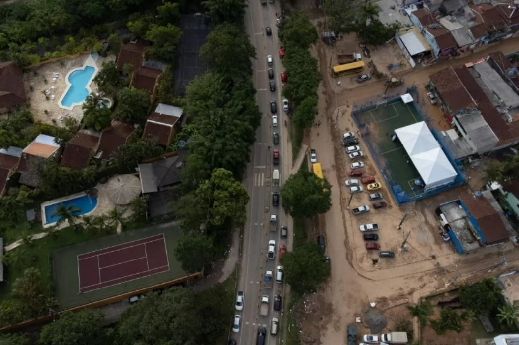 A imagem mostra a rodovia Rio-Santos. De um lado, casas de alto padrão com piscina e rua asfaltada. Do outro, casas simples e com estrada de terra.