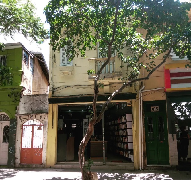 Entrada do Sebo Clepsidra. Casa de dois andares amarela, na entrada se vê vários livros. Na frente da loja existe uma árvore com um tronco torto